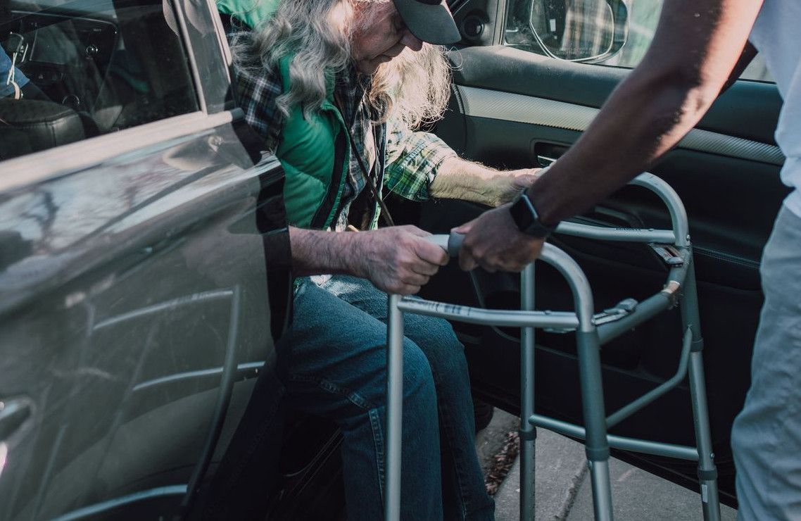 Two men help an elderly person with a walker get out of a car outdoors.